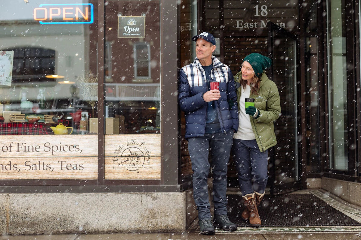 A couple is standing in a cafe entrance holding cups and looking down the snowy street